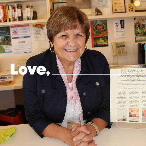 A smiling older woman with short brown hair is wearing a white shirt with a black blazer. She has a pink scarf tied around her neck and is wearing gold hoop earrings. She is leaning on a white counter and there are books on display behind her.